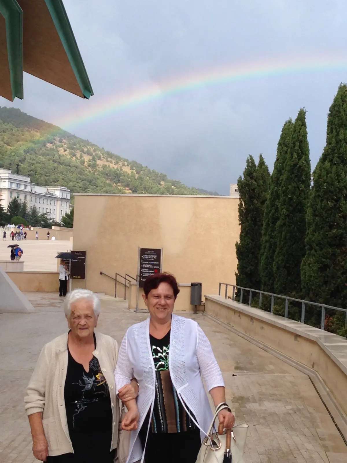Aida, de pelo blanco, y su hija Annamaria, del brazo, en la explanada del santuario; detrás, un muro claro, cipreses, la montaña y un arcoíris que cruza el cielo nublado