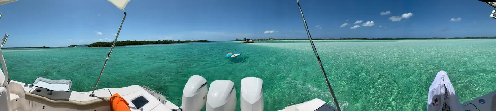 Desde la popa de una lancha, junto a sus tres motores fuera de borda, un agua turquesa tan clara que deja ver el fondo; dos tablas flotan cerca y, al fondo, hay cayos bajos y una barra de arena