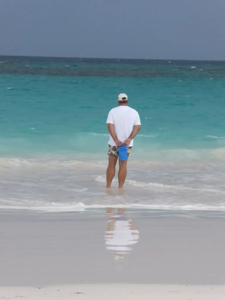Un hombre de espaldas, de pie en la orilla con las manos a la espalda, mira un mar turquesa; al fondo, las olas rompen sobre la línea oscura del arrecife