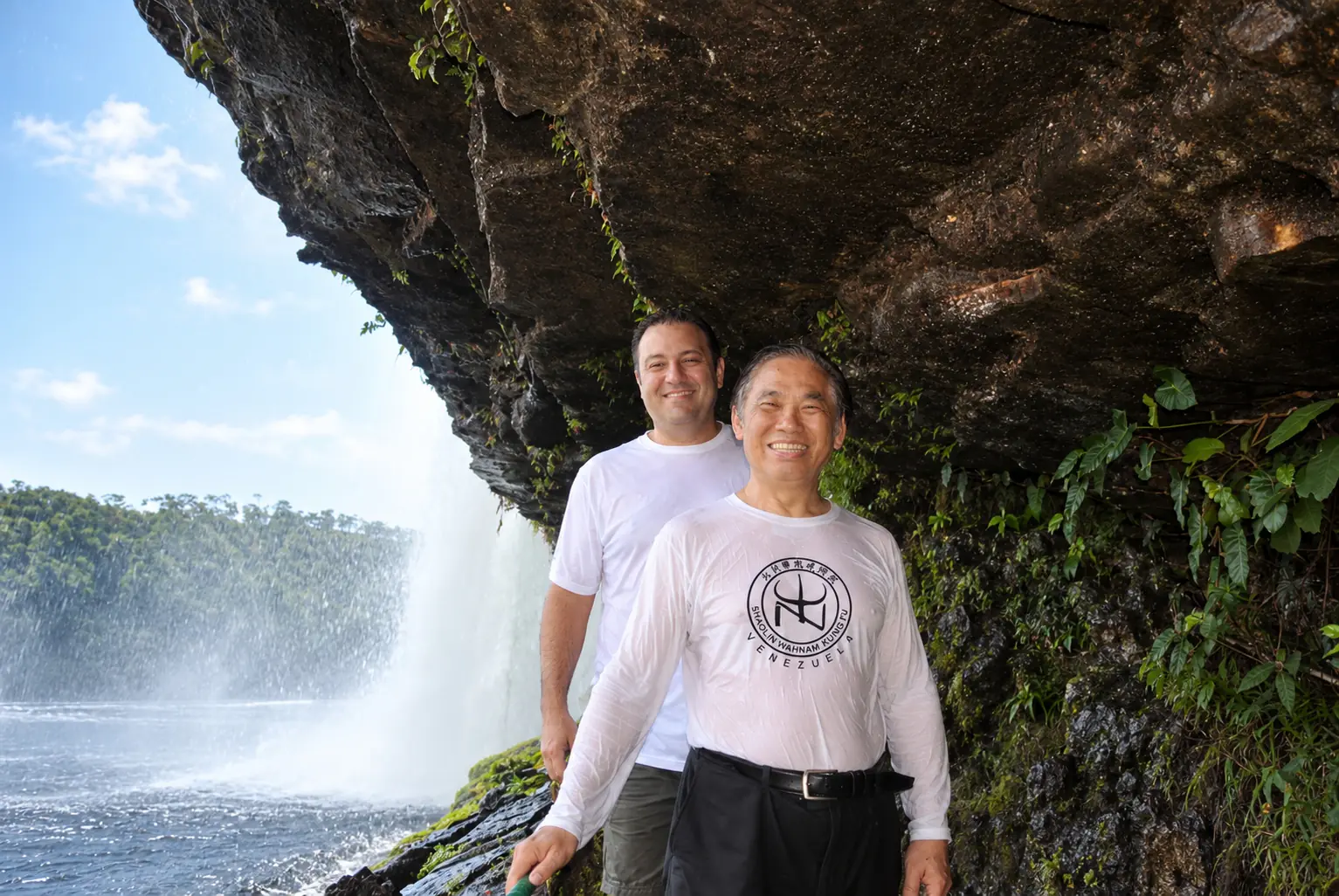 Dos hombres sonríen bajo la roca, detrás de la cortina de agua del salto; delante, Sigung, con la camiseta empapada de Shaolin Wahnam Kung Fu Venezuela; a la izquierda, el agua cae sobre la laguna y, al fondo, la selva